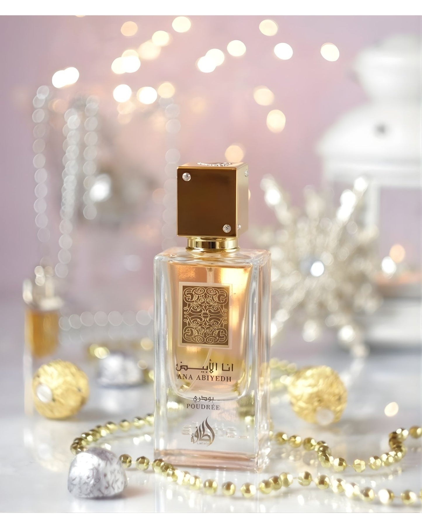 The 'Ana Abiyedh Poudree' perfume bottle, with its gold cap and pale pink liquid, is centered on a surface surrounded by gold and silver beaded garlands and foiled chocolates. The background is a soft, pink, and festive blur of bright bokeh lights and a white decorative lantern.