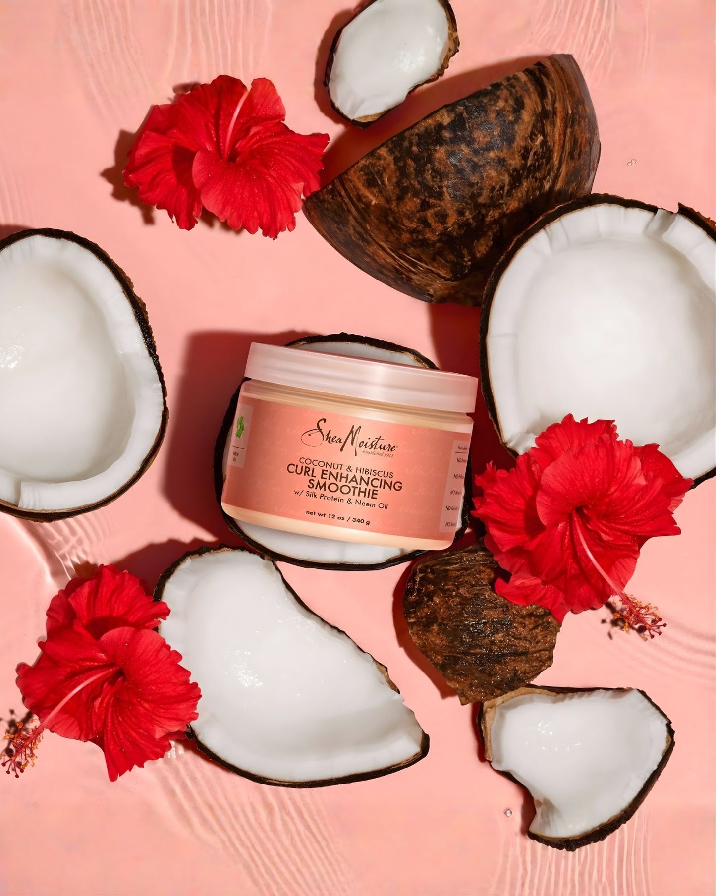 A jar of Shea Moisture Coconut & Hibiscus Curl Enhancing Smoothie arranged on a pink surface with fresh coconut halves and red hibiscus flowers.