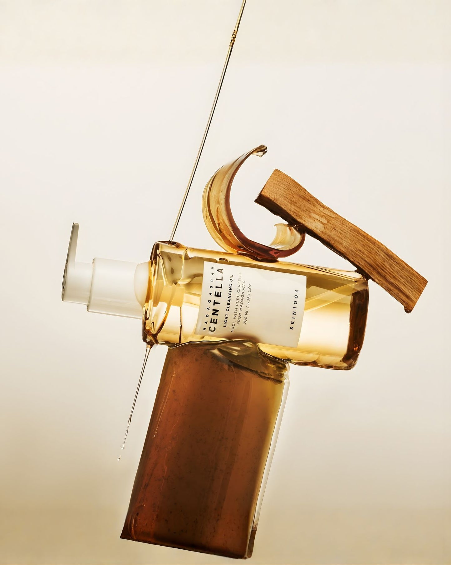 An artistic, stylized product shot of the Skin1004 Madagascar Centella Light Cleansing Oil bottle, positioned horizontally with amber wood shavings and oil dripping across it, conveying a natural aesthetic.