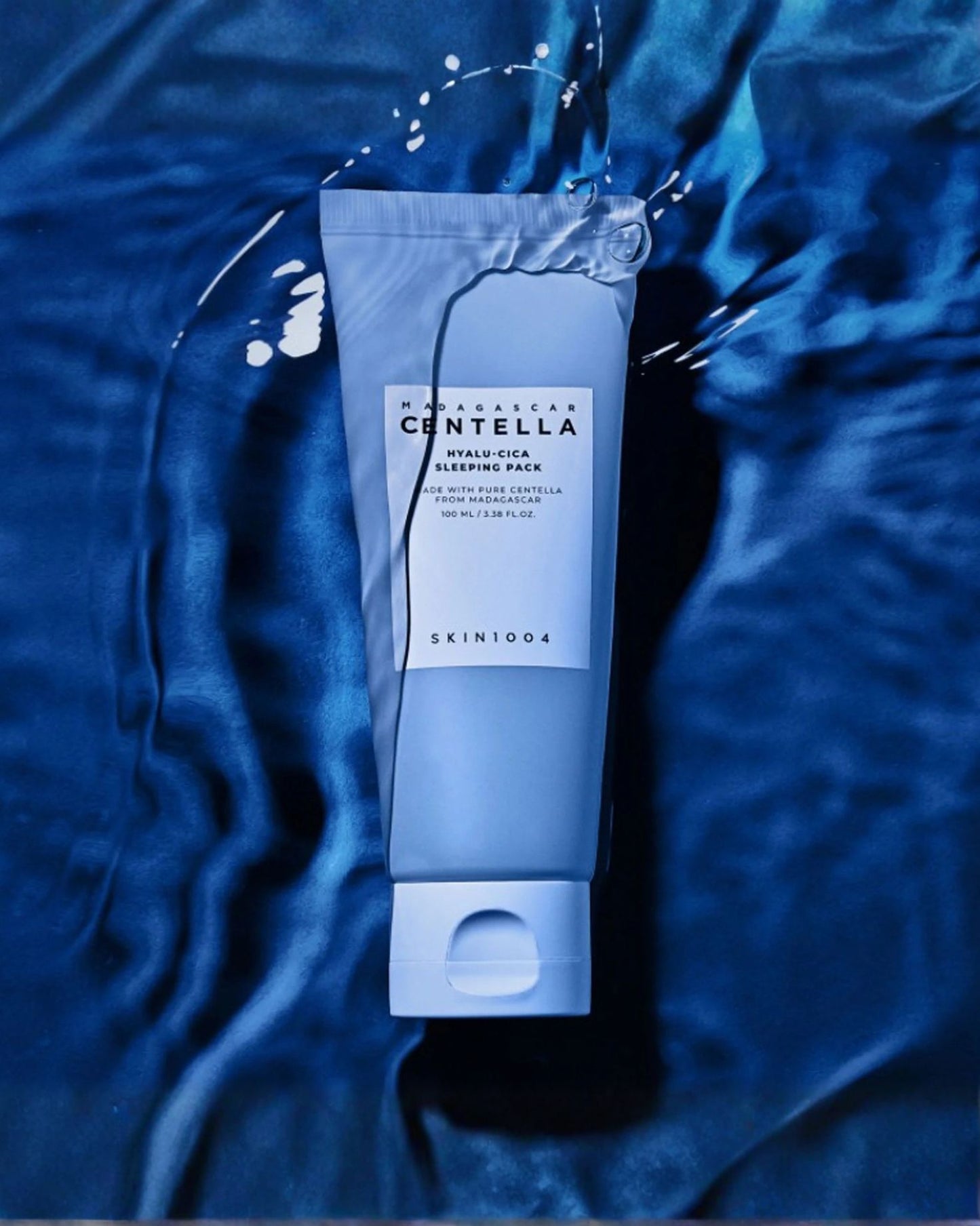 Light blue Hyalu-Cica Sleeping Pack tube resting on dark blue, silky fabric, with water drops on the packaging.