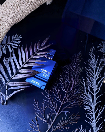 A flat lay photo of the dark blue Al Haramain Amber Oud Dubai Night EXdp perfume bottle, surrounded by metallic silver-blue painted fern leaves and branches. A piece of textured cream fabric is visible in the top left corner, all set on a reflective dark blue surface.