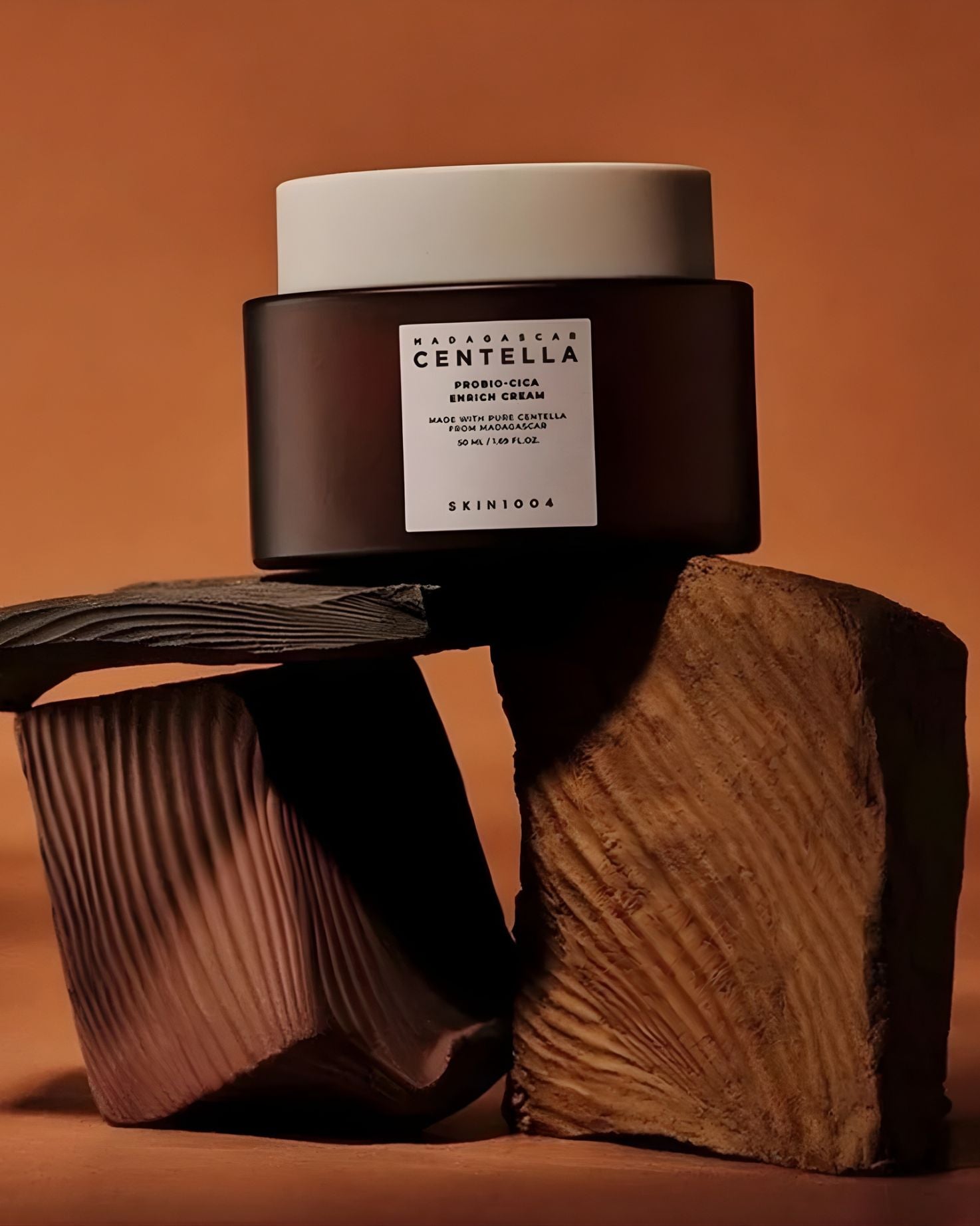 SKIN1004 Madagascar Centella Probio-Cica Enrich Cream jar resting on stacked, textured wooden blocks with an orange-brown background, creating an earthy product photo.