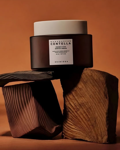 SKIN1004 Madagascar Centella Probio-Cica Enrich Cream jar resting on stacked, textured wooden blocks with an orange-brown background, creating an earthy product photo.