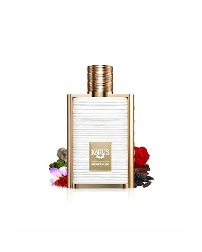 The gold-framed Karus Secret Musk perfume bottle with a textured white center, set amidst various scent ingredients, including a red rose, saffron threads, cinnamon sticks, a purple flower, and dark seeds, against a white background.