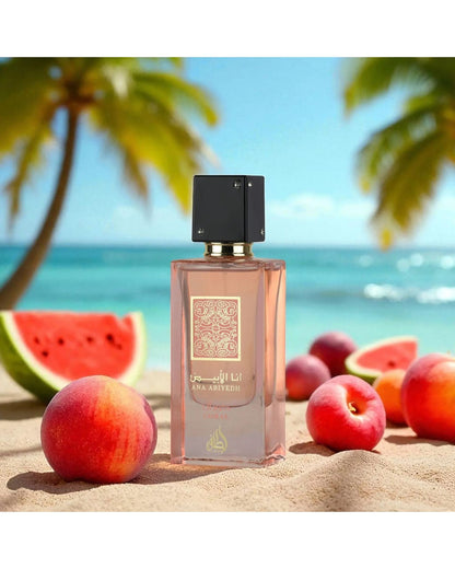 The Ana Abiyedh Coral perfume bottle is set on a sandy beach, surrounded by whole peaches and slices of watermelon. The background features a turquoise ocean, a clear blue sky, and blurred palm trees, emphasizing a tropical, fruity theme.