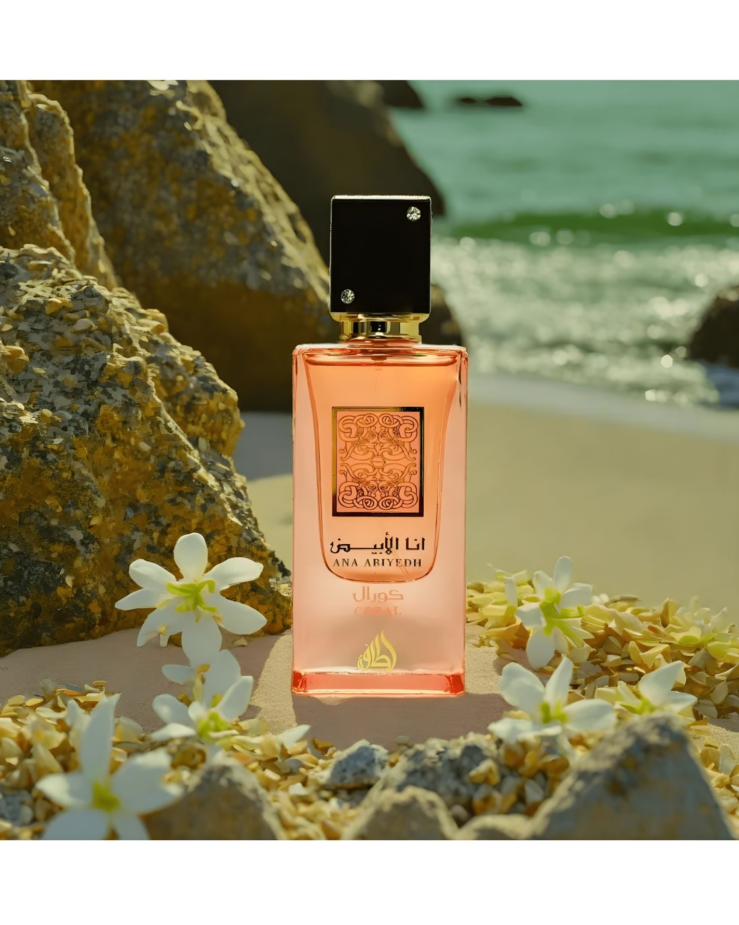 The Ana Abiyedh Coral perfume bottle sits on a sandy shore decorated with small white flowers and dried petals. Large, tan rocks frame the left, and the turquoise ocean and light waves are visible in the background, suggesting a serene coastal setting.