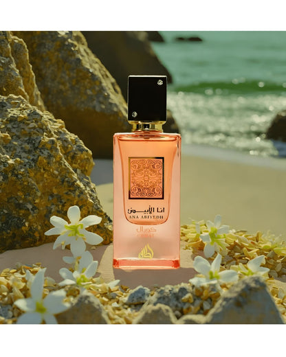 The Ana Abiyedh Coral perfume bottle sits on a sandy shore decorated with small white flowers and dried petals. Large, tan rocks frame the left, and the turquoise ocean and light waves are visible in the background, suggesting a serene coastal setting.