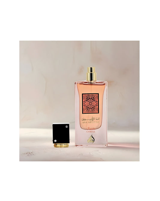 A rectangular perfume bottle with pink liquid that gradients to clear glass at the base. The front label is coral pink with ornate dark brown Arabic calligraphy and the text 'ANA ABIYYEDH CORAL'. The black and gold cap sits detached beside the bottle on a light, marble-like surface.
