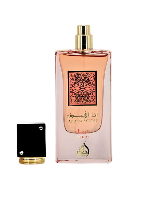 A rectangular perfume bottle with pink liquid that gradients to clear glass at the base. The front label is coral pink with ornate dark brown Arabic calligraphy and the text 'ANA ABIYYEDH CORAL'. The black and gold cap sits detached beside the bottle on a light, marble-like surface.