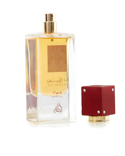 A clear, rectangular perfume bottle containing yellow/amber liquid, labeled with gold Arabic calligraphy and the text ANA ABIYEDH ROUGE Edp. The detached cap is a cubic, matte red with gold accents and four small clear jewels.