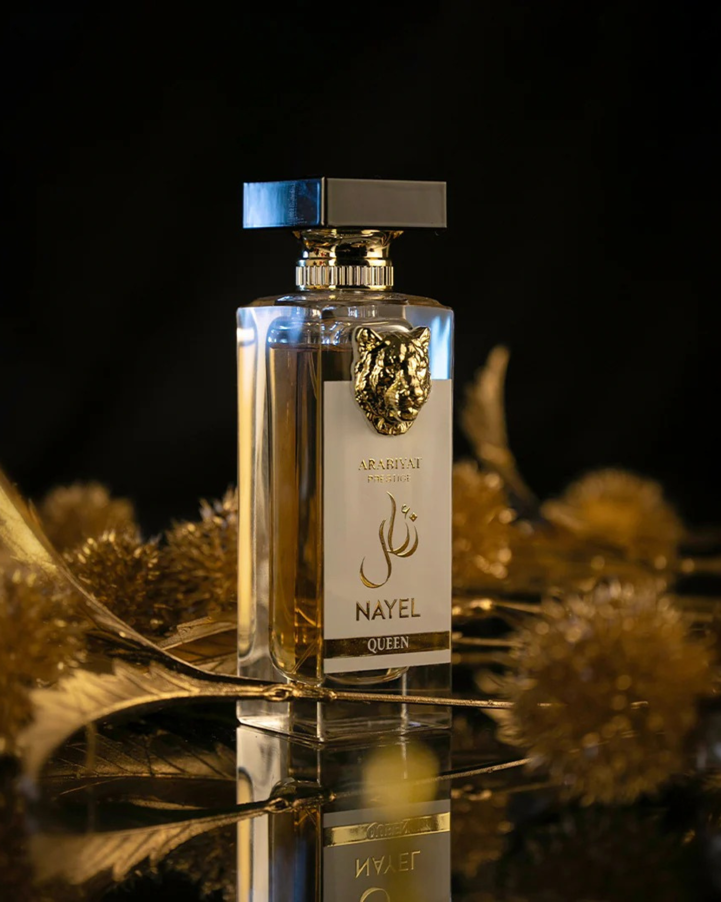 The Arabiyat Prestige 'Nayel Queen' perfume bottle, featuring a prominent gold lion's head emblem and a black and gold cap, is displayed on a reflective dark surface. It is surrounded by blurred, gilded leaves and dark gold ornamental elements, against a dramatic black background.