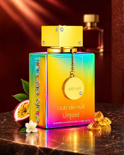 Armaf Club de Nuit Untold Eau de Parfum bottle in an iridescent rainbow gradient, displayed with a sliced passion fruit, small white flower, and amber crystals on a marble surface.