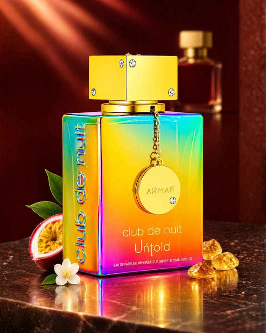 Armaf Club de Nuit Untold Eau de Parfum bottle in an iridescent rainbow gradient, displayed with a sliced passion fruit, small white flower, and amber crystals on a marble surface.