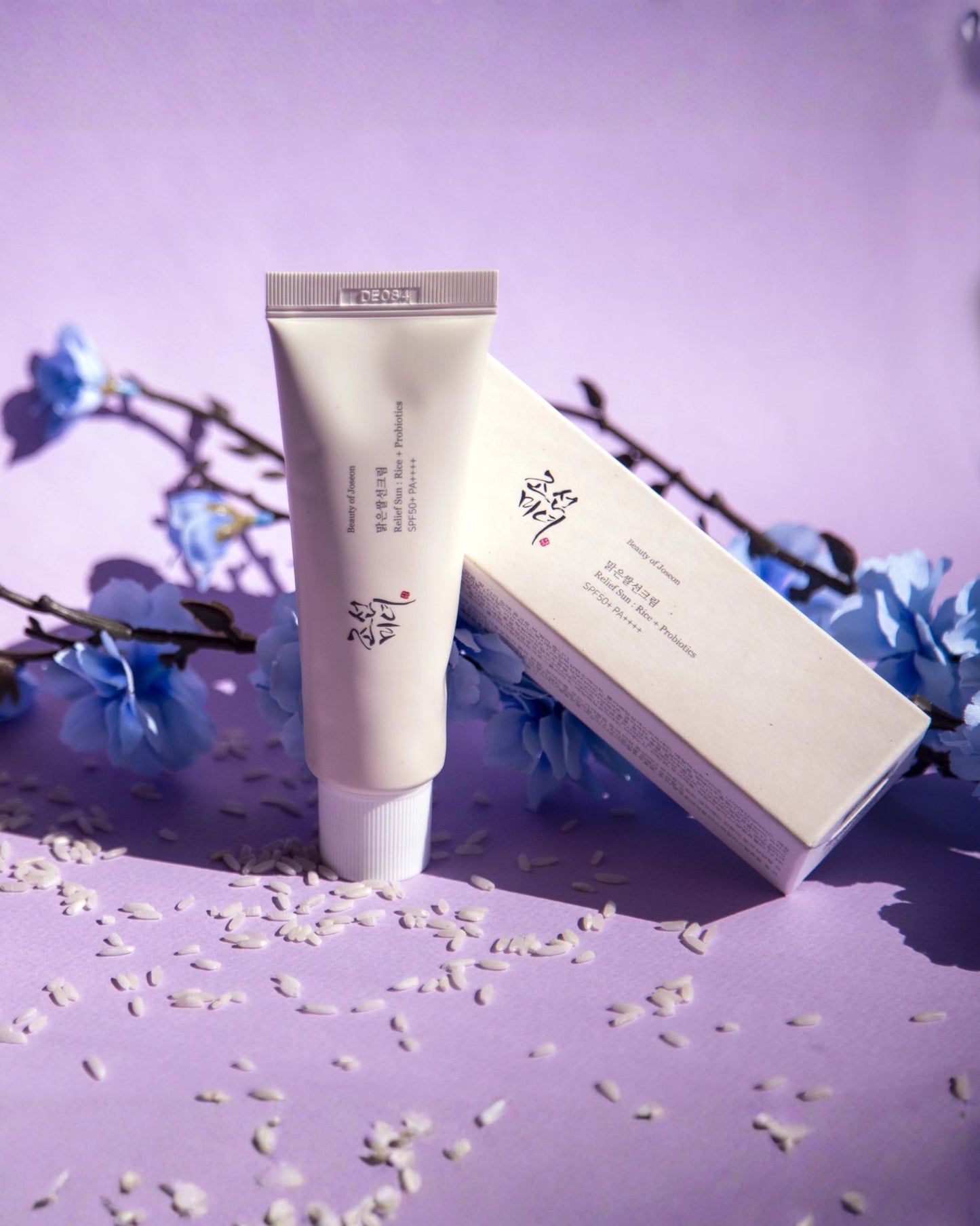 Beauty of Joseon Relief Sun: Rice + Probiotics SPF 50+ PA++++ tube and box, set on a lavender background with scattered rice grains and blue flower sprigs for a serene aesthetic.