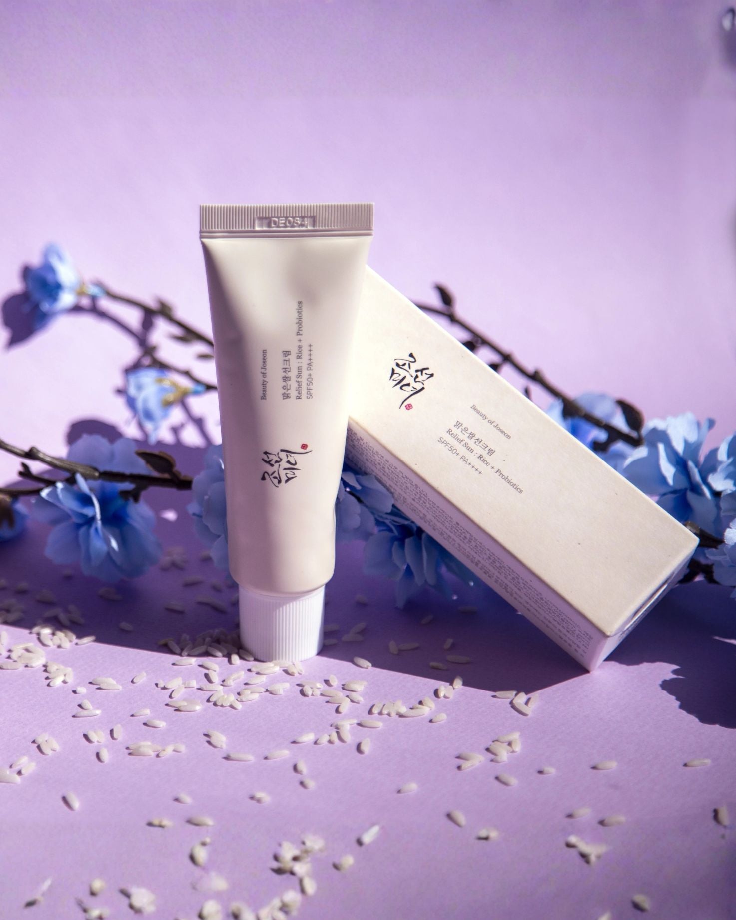 Beauty of Joseon Relief Sun: Rice + Probiotics SPF 50+ PA++++ tube and box, set on a lavender background with scattered rice grains and blue flower sprigs for a serene aesthetic.