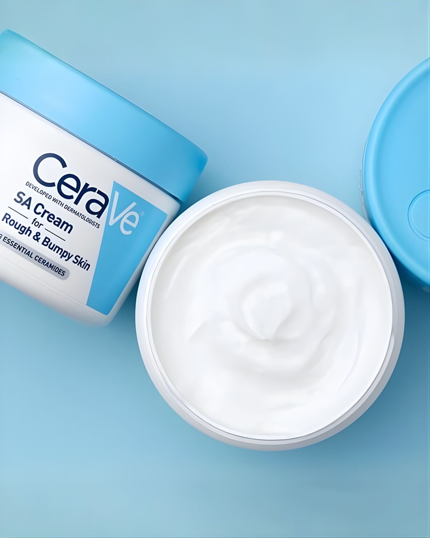An overhead close-up shot of an open tub of CeraVe SA Cream for Rough & Bumpy Skin, showing the thick white cream texture and the blue lid on a light blue background.