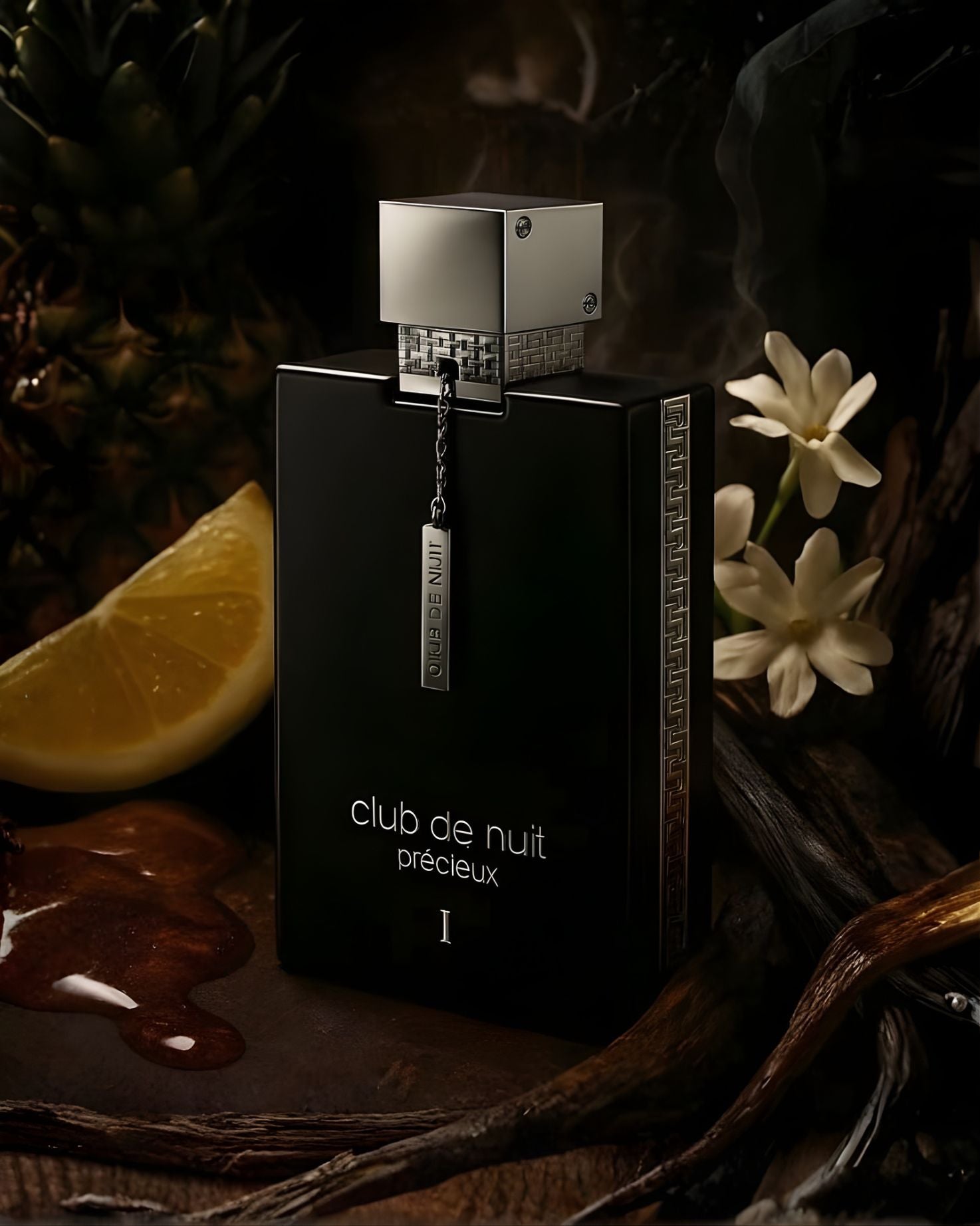 A dramatic photo of the black Club De Nuit Precieux 1 EXdp perfume bottle surrounded by fragrance notes, including a slice of lemon, jasmine flowers, pineapple, vanilla pods, and a dark liquid spill.