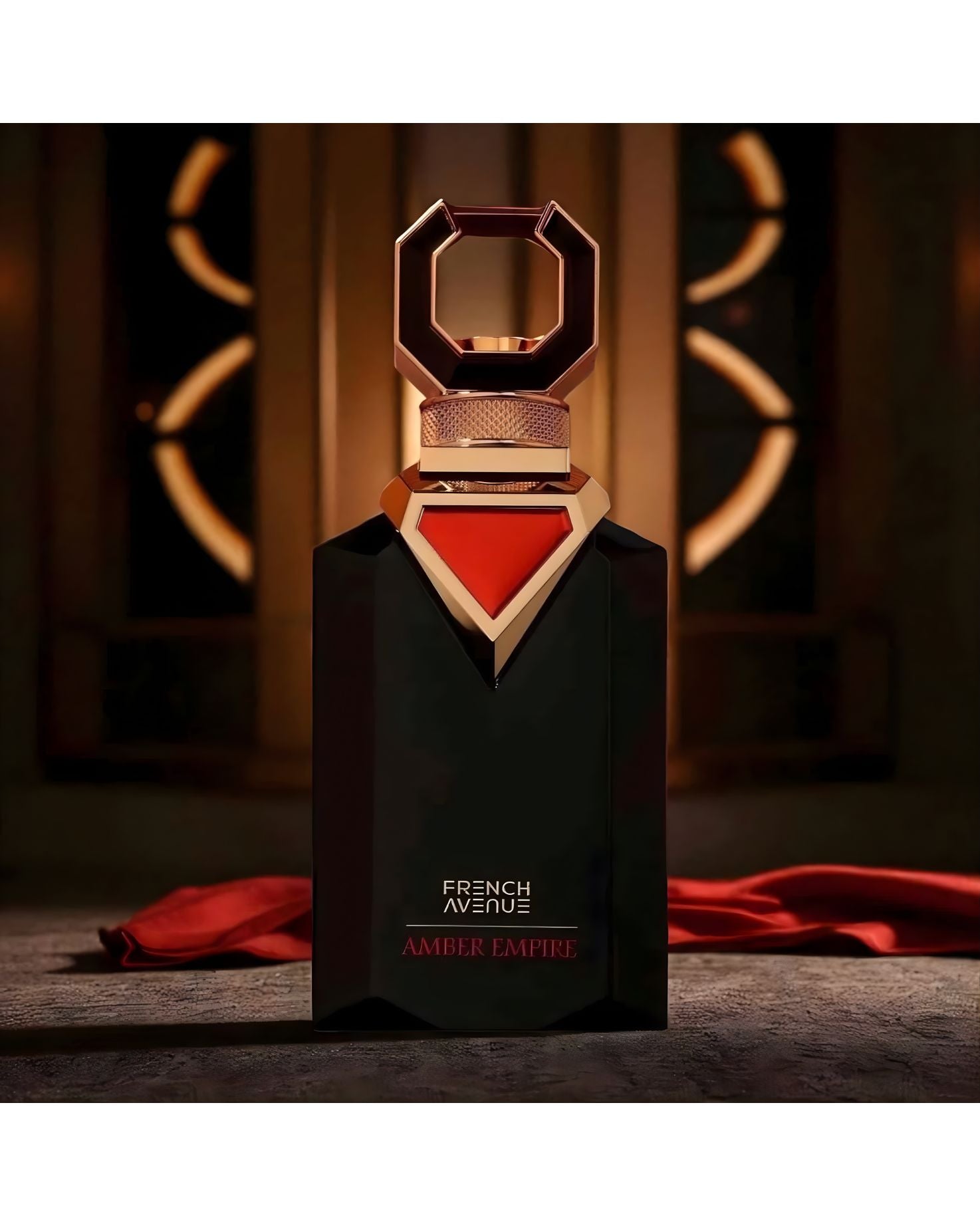 The black, faceted 'French Avenue Amber Empire' perfume bottle, with its bold gold cap and red triangle accent, is displayed on a textured stone surface with a draped piece of red fabric. The background is dark and blurred, featuring ornate, illuminated geometric shapes suggestive of a grand interior.