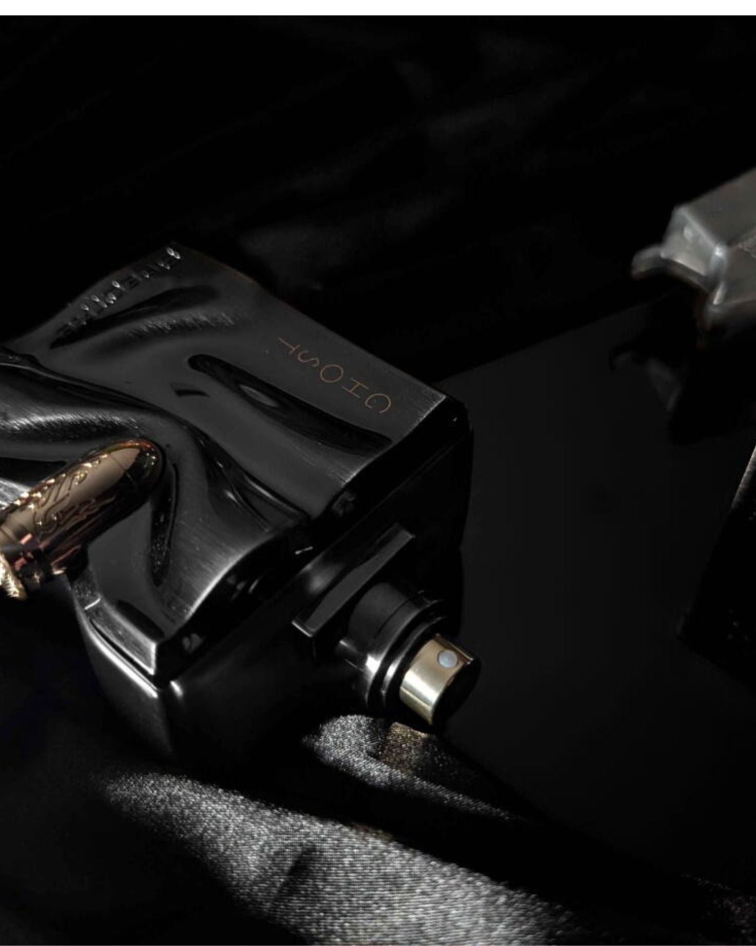 A moody, dark close-up of the black "Ghost Spectre" perfume bottle, lying on dark, satiny fabric. The shot focuses on the gold bullet detail, the "GHOST" text, and the metallic spray nozzle, with a dark, blurred background.