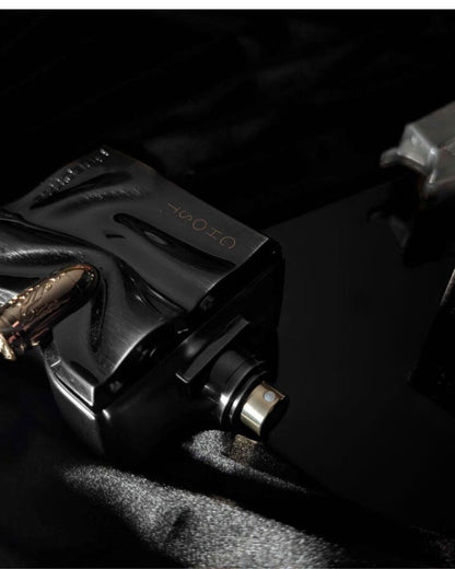 A moody, dark close-up of the black "Ghost Spectre" perfume bottle, lying on dark, satiny fabric. The shot focuses on the gold bullet detail, the "GHOST" text, and the metallic spray nozzle, with a dark, blurred background.