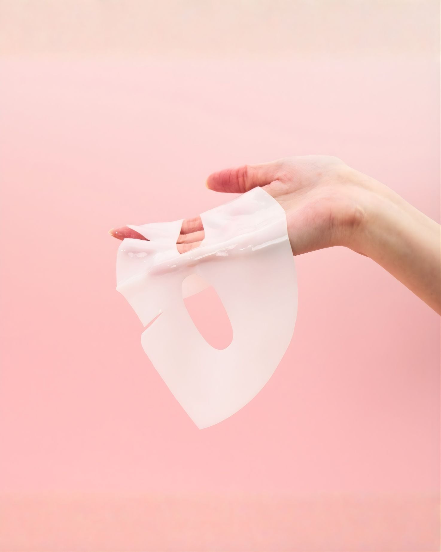 A hand gently holding up a clear, white essence-soaked facial sheet mask against a soft pink background. The mask is transparent and appears highly moisturizing.