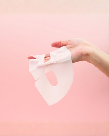 A hand gently holding up a clear, white essence-soaked facial sheet mask against a soft pink background. The mask is transparent and appears highly moisturizing.