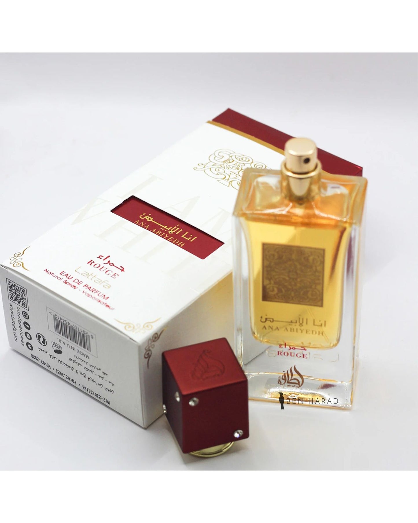 The Lattafa ANA ABIYEDH ROUGE Edp perfume bottle, with its amber liquid and gold accents, stands next to its open white and red retail box. The bottle's detached red cubic cap is in the foreground. All items are displayed on a white surface.