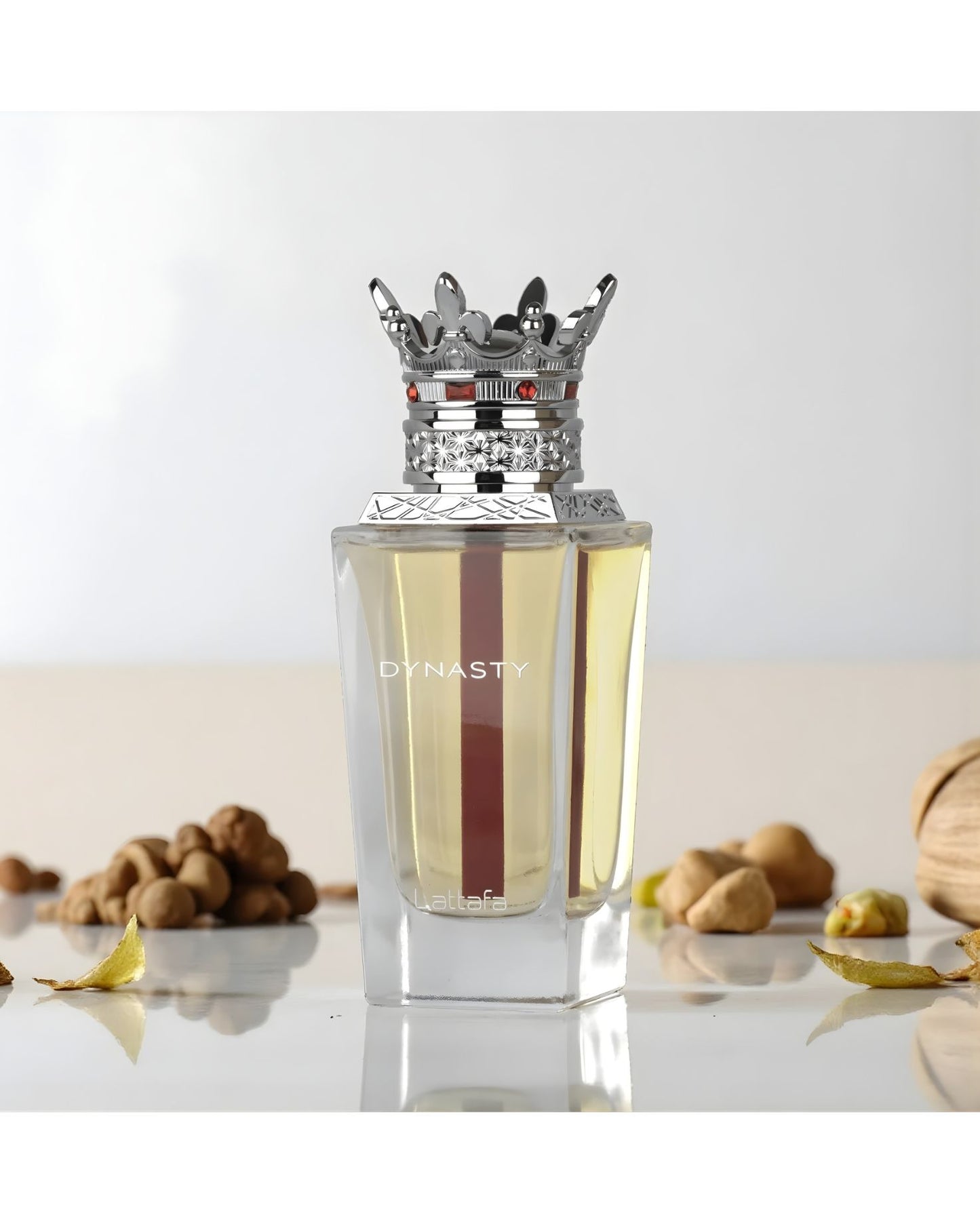The Lattafa 'Dynasty' EDP perfume bottle, with its clear glass, yellow liquid, red stripe, and elaborate silver crown cap, is displayed on a light surface surrounded by various nuts, dried leaves, and a textured, woven object. The background is soft and bright white.