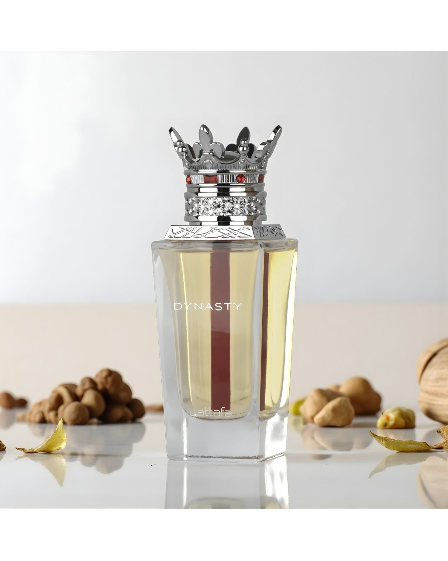 The Lattafa 'Dynasty' EDP perfume bottle, with its clear glass, yellow liquid, red stripe, and elaborate silver crown cap, is displayed on a light surface surrounded by various nuts, dried leaves, and a textured, woven object. The background is soft and bright white.