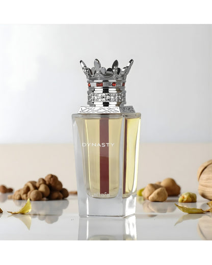 The Lattafa 'Dynasty' EDP perfume bottle, with its clear glass, yellow liquid, red stripe, and elaborate silver crown cap, is displayed on a light surface surrounded by various nuts, dried leaves, and a textured, woven object. The background is soft and bright white.