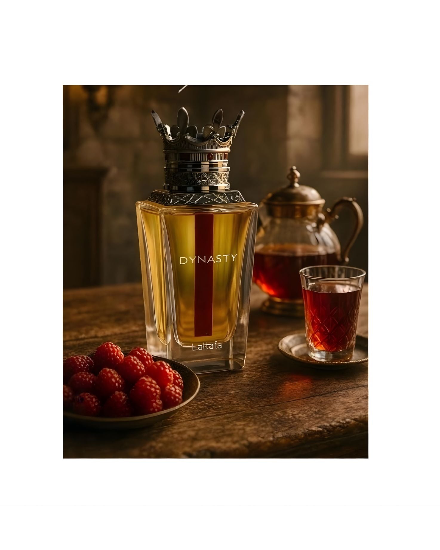The Lattafa 'Dynasty' EDP perfume bottle, with its yellow liquid, red stripe, and ornate crown cap, is placed on a rustic wooden table next to a small bowl of fresh raspberries and a glass of red tea. A decorative teapot is visible in the blurred background.