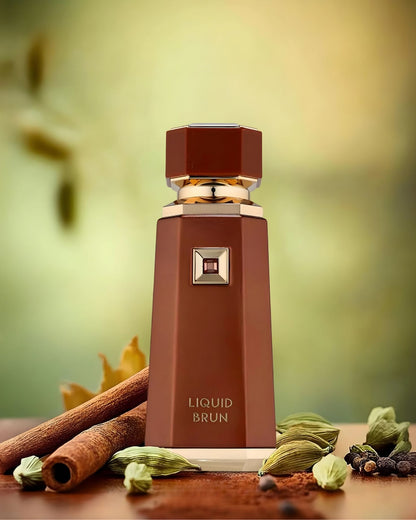 The Liquid Brun perfume bottle, in brown and gold, is presented on a wooden surface surrounded by spices, including cinnamon sticks, green cardamom pods, black peppercorns, and cocoa powder. The background is a soft, blurred green and yellow.
