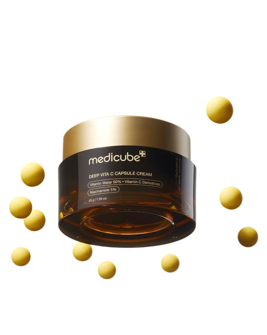 Medicube Deep Vita C Capsule Cream jar, a vitamin-infused face cream, with several bright yellow vitamin capsules scattered around it on a white background.