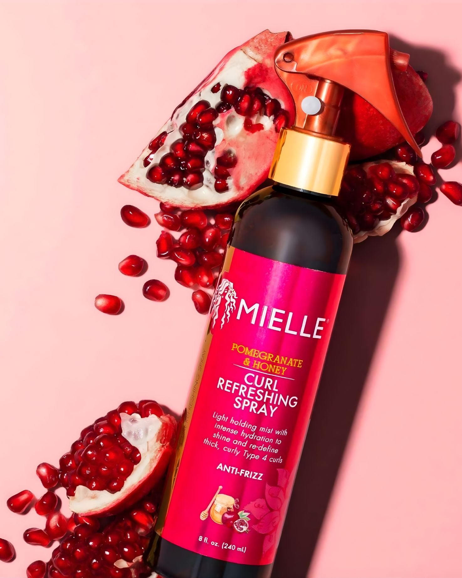 An overhead view of a Mielle Pomegranate & Honey Curl Refreshing Spray bottle on a pink background, surrounded by halved pomegranates and loose pomegranate seeds.