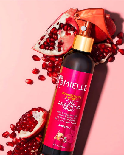 An overhead view of a Mielle Pomegranate & Honey Curl Refreshing Spray bottle on a pink background, surrounded by halved pomegranates and loose pomegranate seeds.