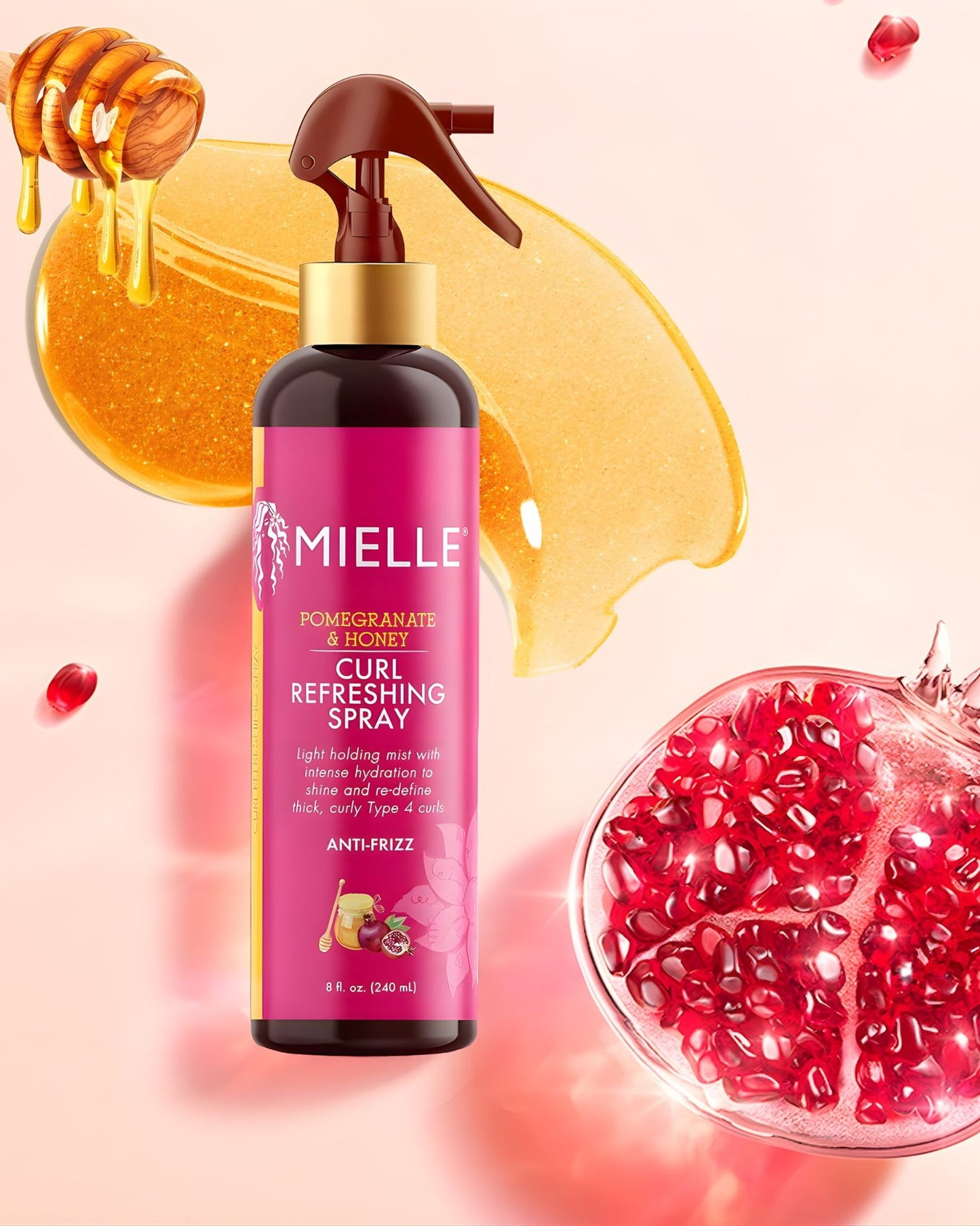 A bottle of Mielle Pomegranate & Honey Curl Refreshing Spray set against a background featuring a large cut pomegranate on the right, dripping honey from a honey dipper in the upper left, and a golden honey smear behind the bottle. The bottle is described on the label as a "light holding mist with intense hydration to shine and redefine thick, curly Type 4 curls" and is "ANTI-FRIZZ.