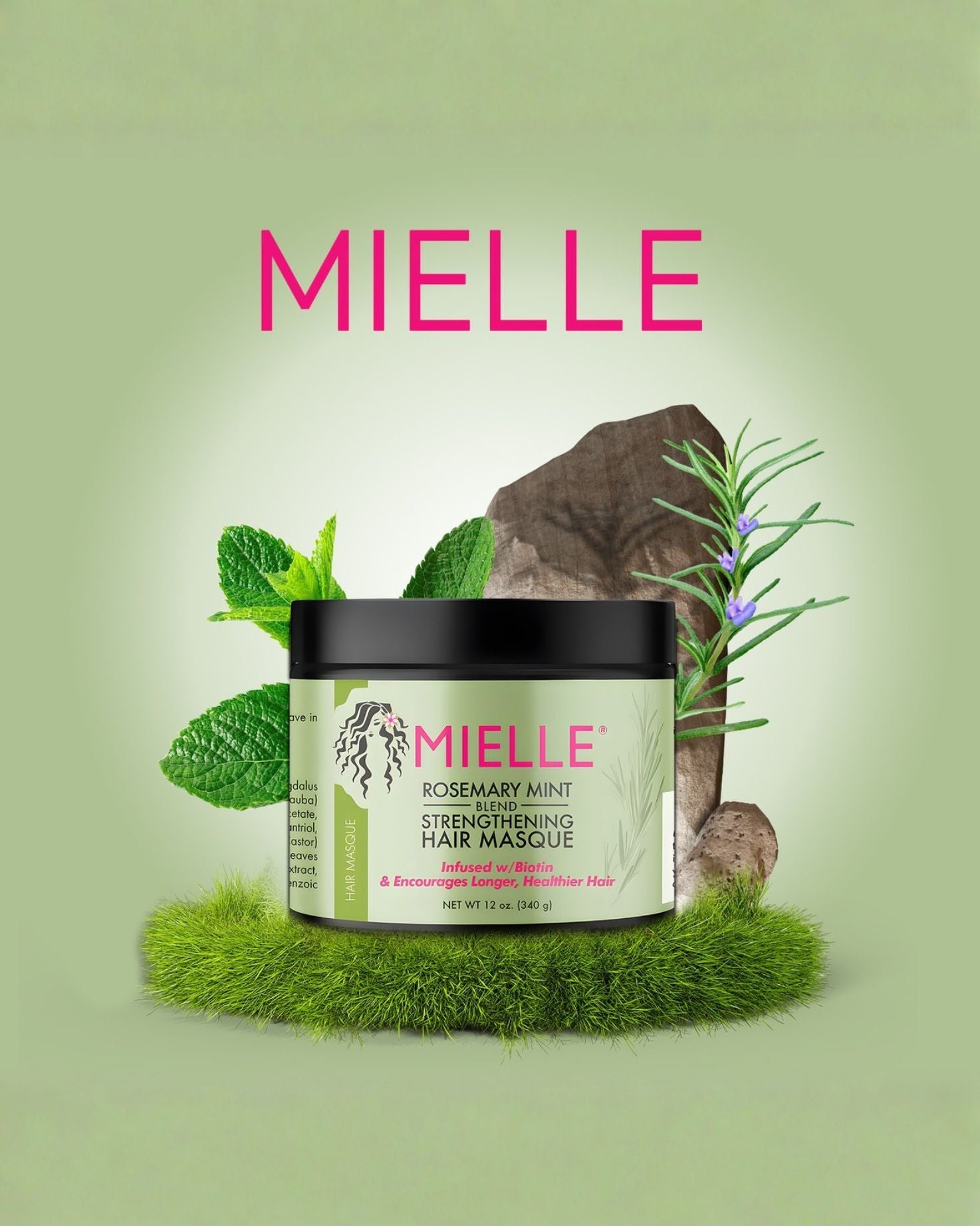 The Mielle Rosemary Mint Strengthening Hair Masque jar is centrally displayed on a bed of bright green artificial moss. It is surrounded by fresh mint leaves, a sprig of flowering rosemary, and pieces of dark wood or rock, set against a background of soft green and pink gradient, with the Mielle logo at the top.