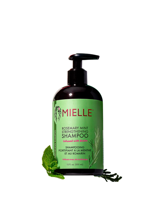 A black pump bottle of Mielle Rosemary Mint Strengthening Shampoo, infused with Biotin. The label is green and white, with sprigs of rosemary and mint illustrating the ingredients. A fresh mint leaf and a rosemary sprig are placed near the base of the bottle, isolated against a white background.