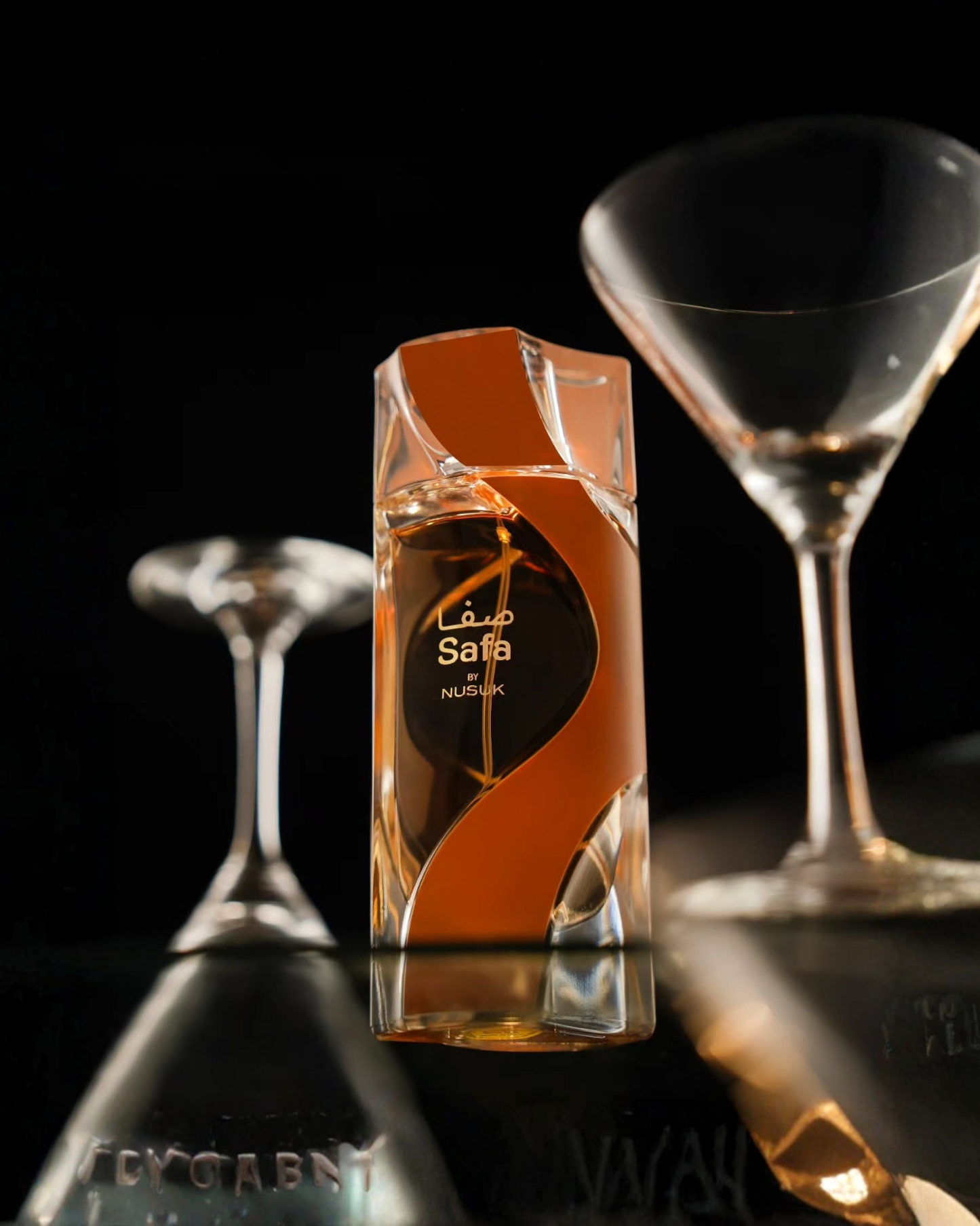 The Nusuk Safa Edpperfume bottle, with its amber liquid and vertical orange/gold swirl design, is centered on a dark, reflective surface. It is flanked by the bases of two inverted clear martini-style glasses, creating a sophisticated, dark, and intimate atmosphere.