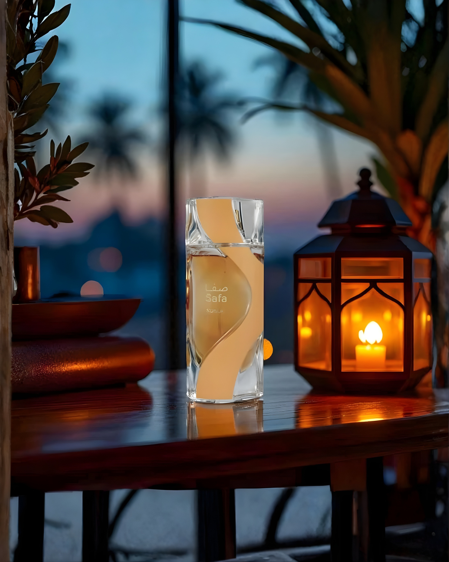 The Nusuk Safa Edp perfume bottle, with its faceted glass and gold-swirled design, is centered on a polished wooden table. It is flanked by a lit, warm-toned lantern on the right, and a dark plant on the left. The background shows a blurred twilight view with palm trees and soft city lights.