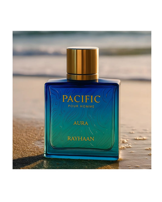 Rayhaan Pacific Aura Pour Homme perfume bottle on a sandy beach with the ocean in the background, featuring a blue and green gradient glass and a gold cap.