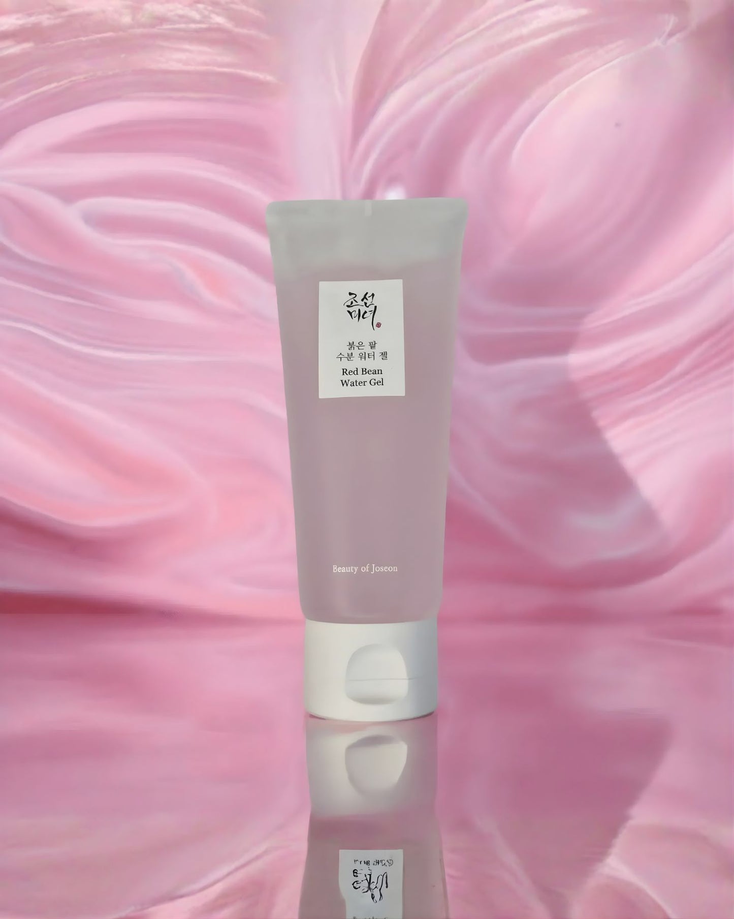 A translucent pink tube of Beauty of Joseon Red Bean Water Gel standing upright on a reflective surface against a swirled, pink, watery background, emphasizing its moisturizing texture