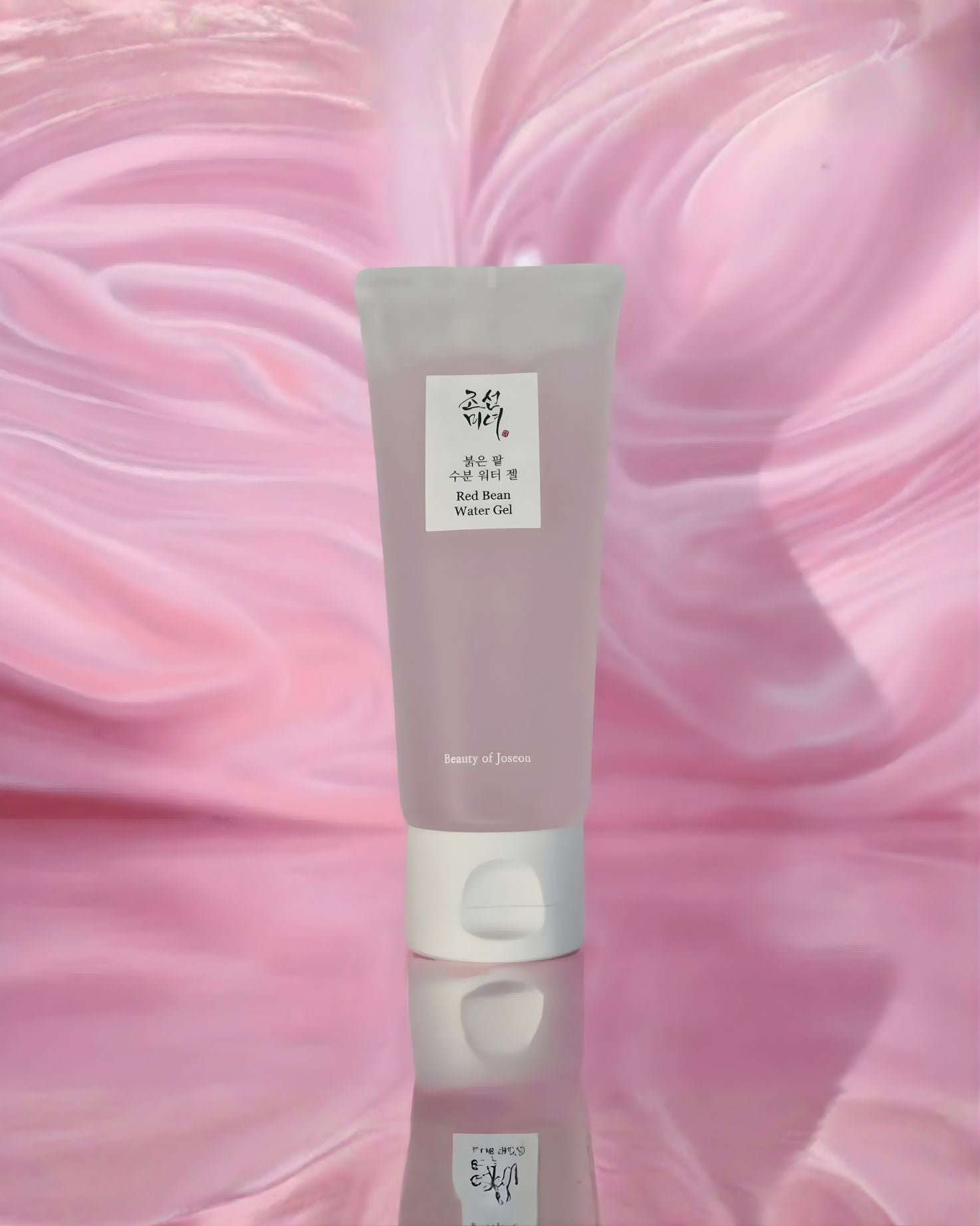 A translucent pink tube of Beauty of Joseon Red Bean Water Gel standing upright on a reflective surface against a swirled, pink, watery background, emphasizing its moisturizing texture
