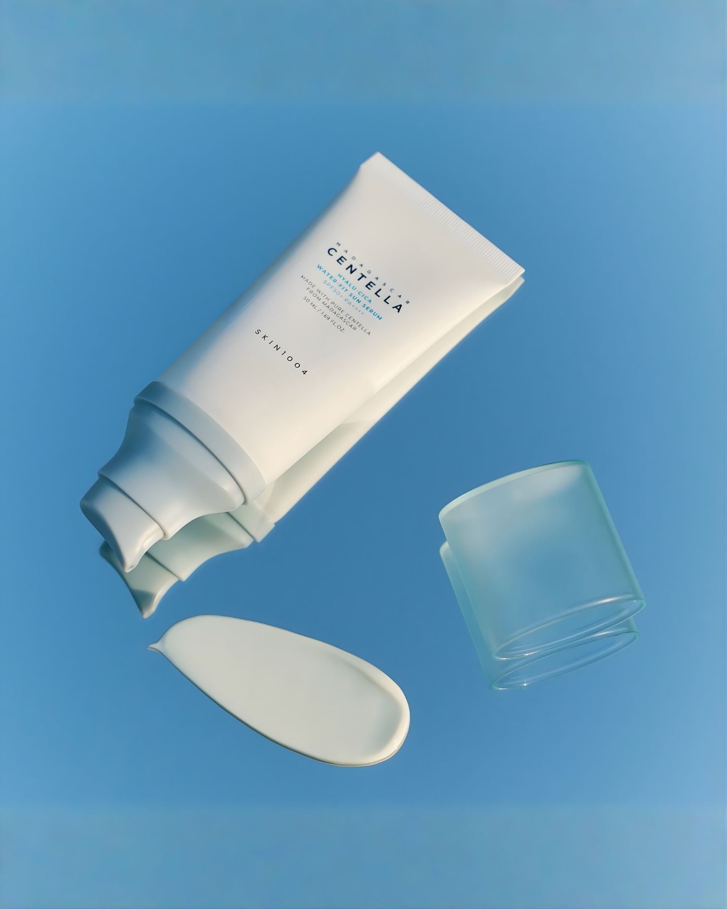 SKIN1004 Madagascar Centella Water-Fit Sun Serum in a white tube, with a swatch of the product on a reflective, light blue surface next to the clear cap.