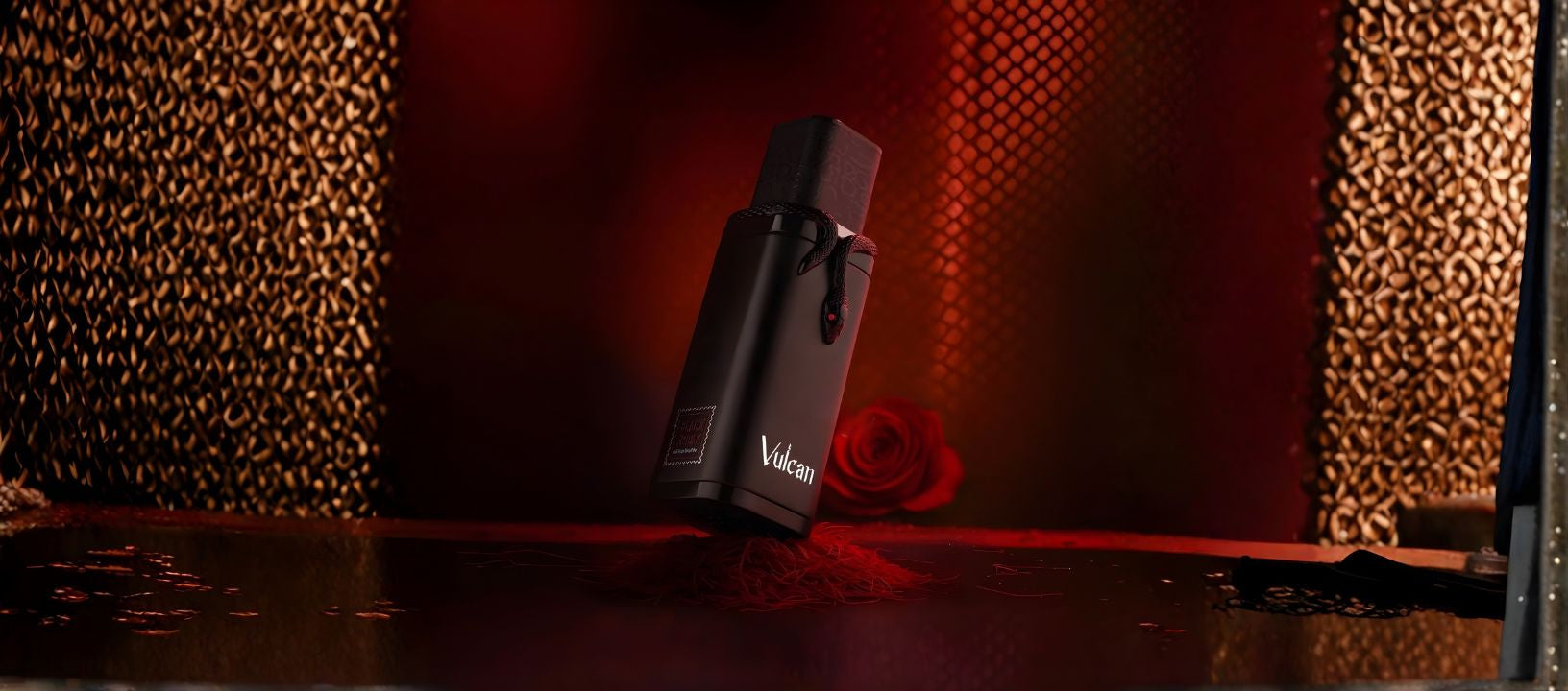 A dramatic, dark lifestyle photo of a small, matte black, rectangular product bottle labeled "Vulcan," possibly a perfume or cosmetic. The bottle has a black cap and appears to be sitting on a metallic surface with dark red threads, a blurred red rose, and a textured gold/dark background.