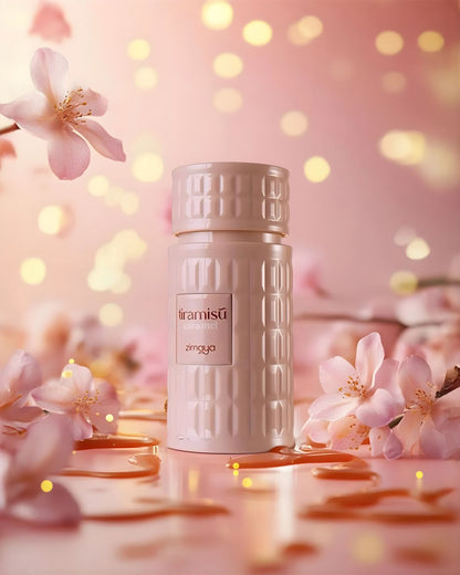 Soft-focus image of a textured, light pink Zimaya 'Tiramisû Caramel' perfume bottle surrounded by delicate pink cherry blossom flowers. The foreground is glossy with scattered liquid droplets and golden light reflections.
