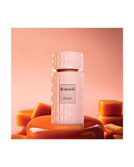 Pink, textured perfume bottle labeled 'Tiramisû' by Zimaya, resting on a glossy caramel-colored pedestal, surrounded by out-of-focus caramel candies on an orange-pink background.